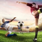 Jugadores de fútbol en acción durante un partido, uno en el suelo intentando interceptar el balón mientras el otro ejecuta un tiro. El fondo muestra un estadio iluminado con espectadores.