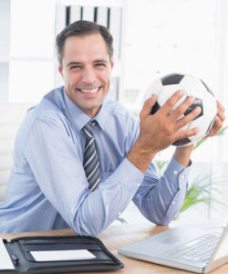 Hombre de negocios sonriente en camisa y corbata sosteniendo un balón de fútbol en una oficina moderna.
