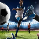 Jugador de fútbol en acción, pateando un balón hacia la portería, mientras el portero se prepara para detenerlo. En el fondo, un estadio iluminado con otros jugadores en el campo.