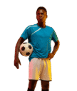 Joven futbolista de pie con una pelota de fútbol en la mano, vistiendo camiseta azul y pantalones cortos blancos, mostrando confianza y actitud deportiva.