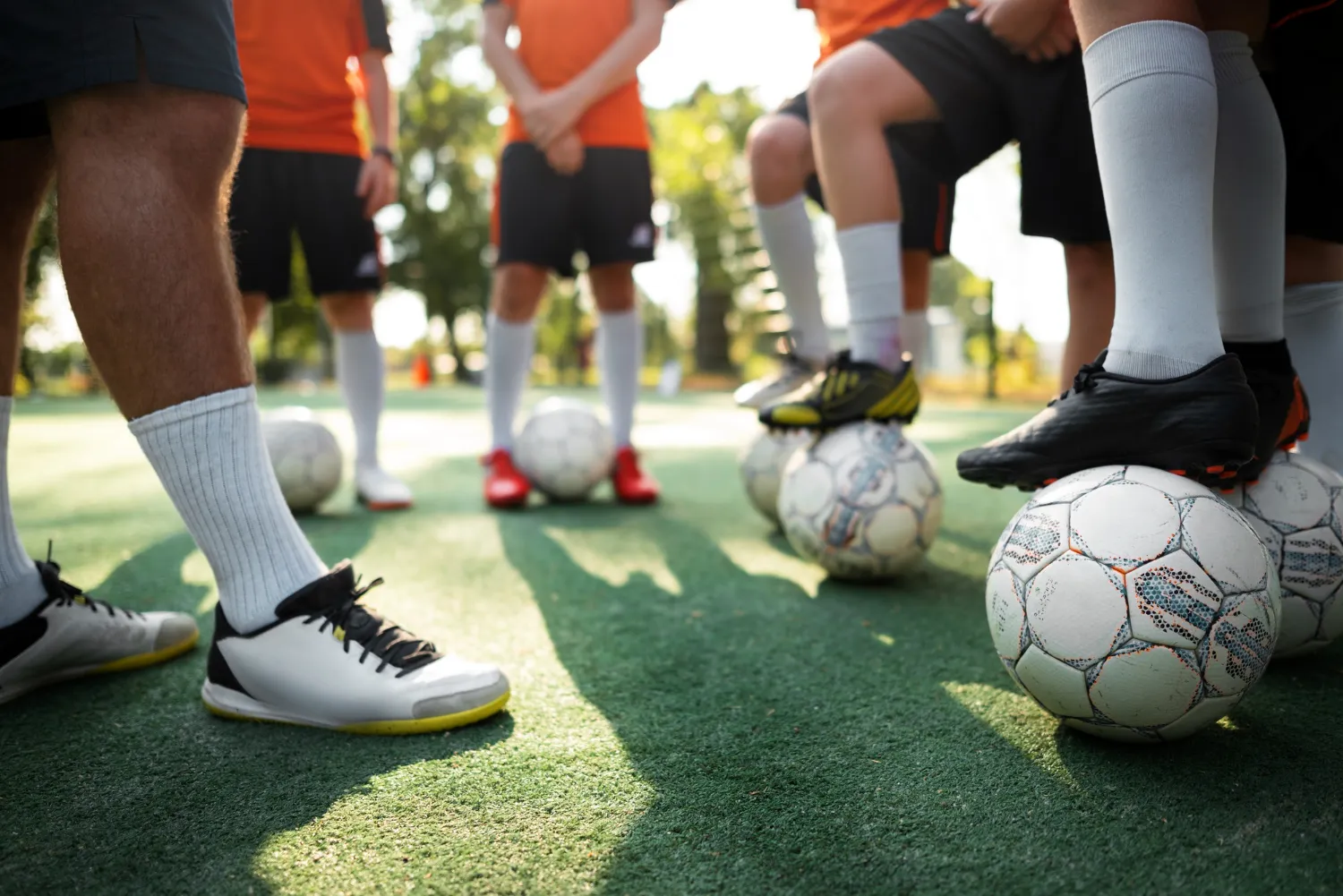 Jugadores de fútbol en una cancha de césped sintético, con pies sobre balones de fútbol, mientras se preparan para un entrenamiento. Visten camisetas naranjas y pantalones cortos, con medias blancas.